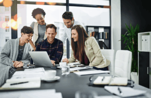 Becoming a Multiskilled Marketer: Resources To Help You Grow A group of marketers in a modern office collaborate around a laptop, smiling as they discuss ideas during a professional training session.