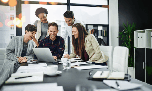 A group of marketers in a modern office collaborate around a laptop, smiling as they discuss ideas during a professional training session.