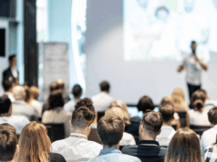 Your Marketing Event Calendar for 2026: Dates, Trends & Insights A large audience of marketers listens to a speaker presenting onstage at a conference, with a bright screen and modern meeting space in the background.