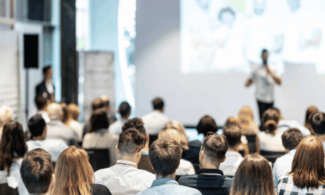 A large audience of marketers listens to a speaker presenting onstage at a conference, with a bright screen and modern meeting space in the background.